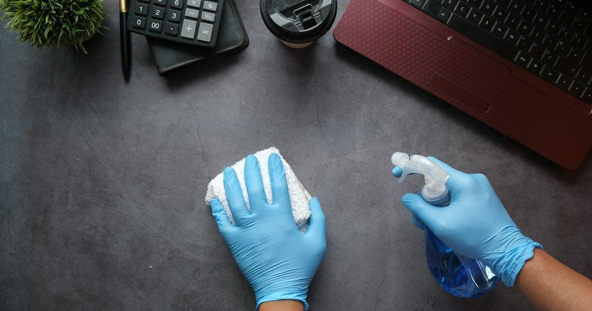 Overhead view of blue-gloved hands wiping a dark desk with a cloth and a blue spray bottle