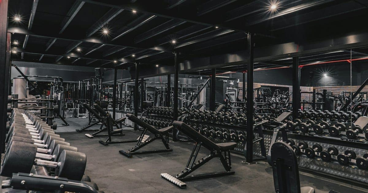 Interno buio di una palestra con rastrelliere di manubri, panche e macchinari sotto faretti a soffitto