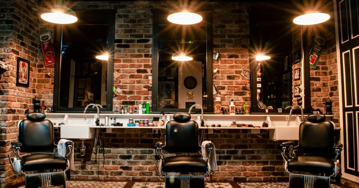 Innenraum eines Barbershops mit drei schwarzen Ledersesseln vor Spiegeln an einer Ziegelwand