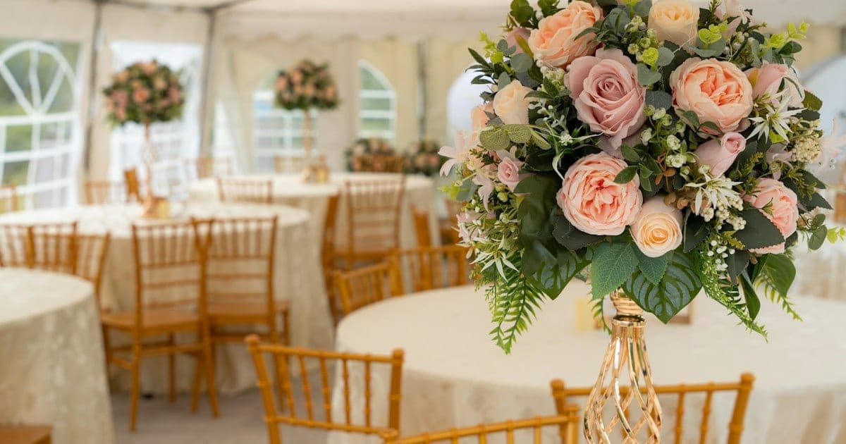 Wedding marquee with round cream-clothed tables, gold chairs and a tall pink rose centrepiece
