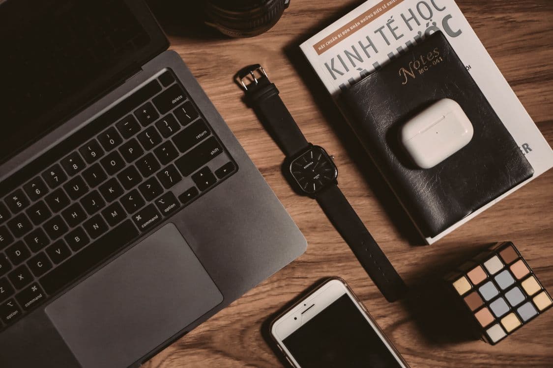 Flat lay of tech gadgets including a laptop, smartphone, book, watch, and AirPods on a wooden table. Photo by Danh Huynh on Pexels