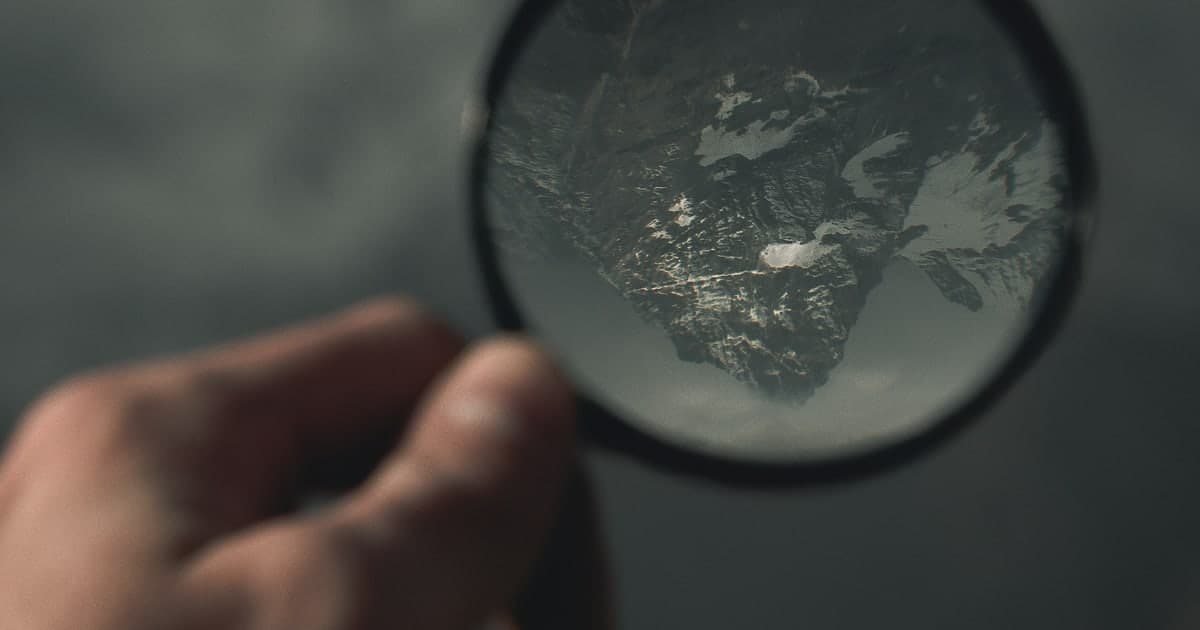 A hand holding a magnifying glass over a map, enlarging a patch of mountain terrain beneath it
