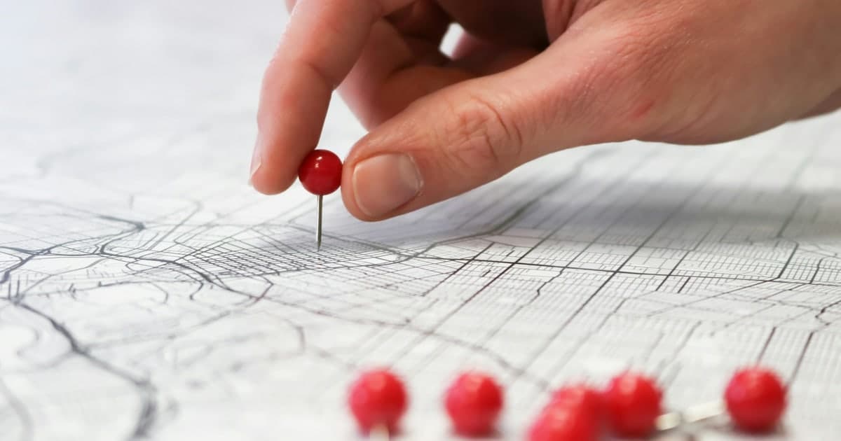 A hand pressing a red pushpin into a printed street map, with more red pins already placed nearby